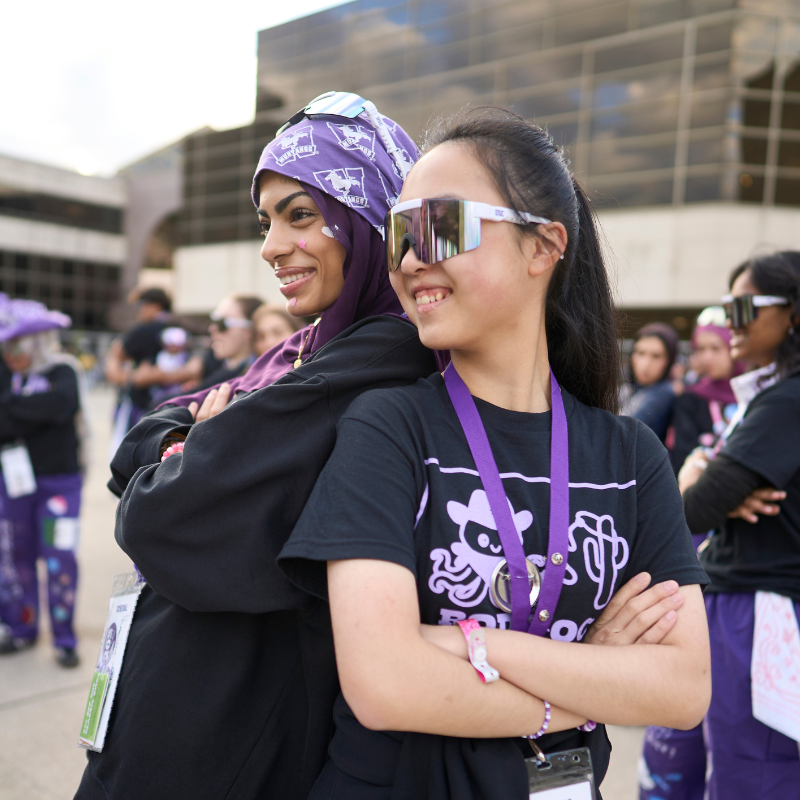 Two young women stand back to back smiling confidently.