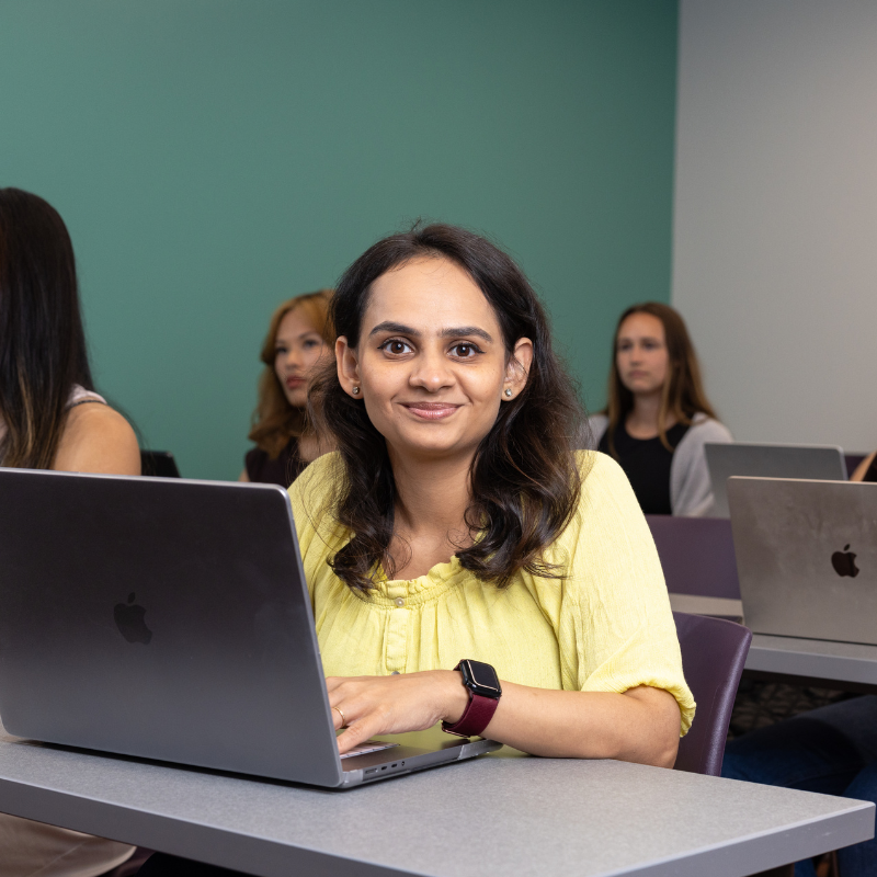 A young woman sits in a classroom setting on a laptop.